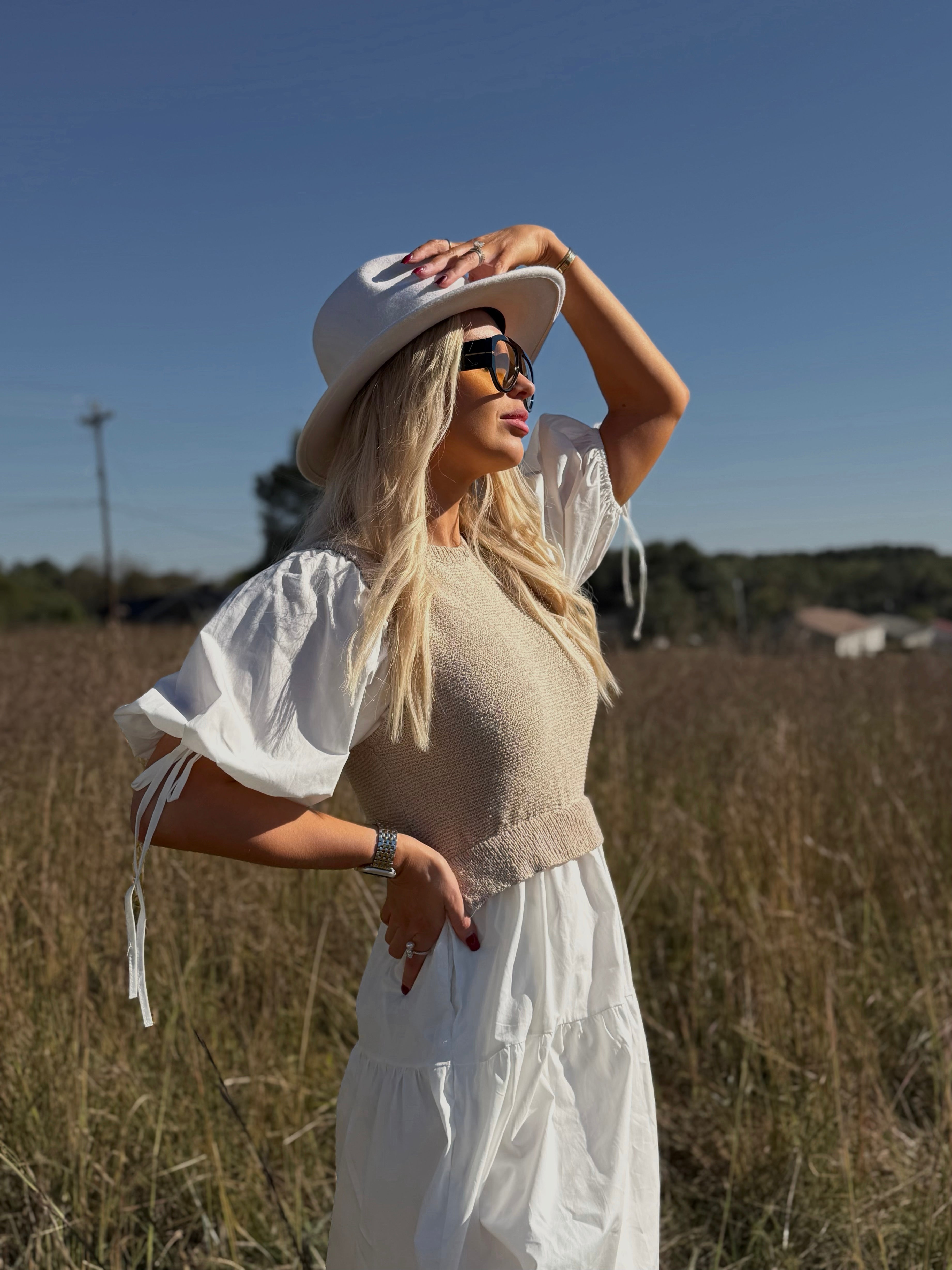 Woman in a white dress and hat standing in a field with a clear blue sky.
